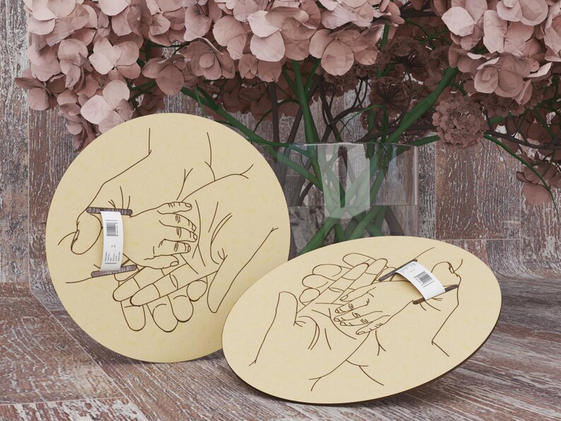 May include: Two light-colored wooden plaques with a hand-drawn illustration of hands holding a baby's hand. One plaque is round, the other is oval. A floral arrangement is in the background.