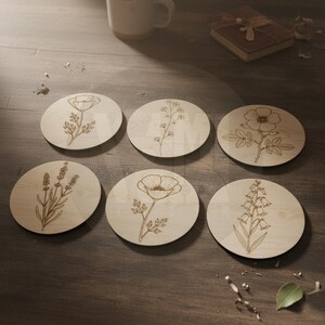Puede incluir: Seis posavasos redondos de madera con dise&ntilde;os florales grabados. Los posavasos presentan varias ilustraciones bot&aacute;nicas, incluyendo una amapola, lavanda y otras flores. Los posavasos est&aacute;n dispuestos sobre una superficie de madera.