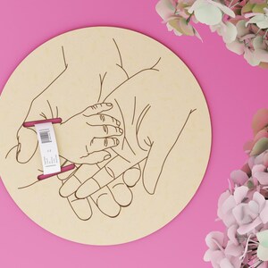 May include: A round wooden plaque with an engraved illustration of three hands, one small hand resting in two larger hands. A tag is attached to the plaque. The background is pink, with flowers in the upper right corner.