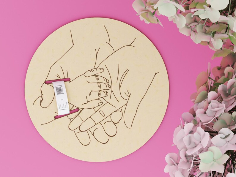May include: A round wooden plaque with an engraved illustration of three hands, one small hand resting in two larger hands. A tag is attached to the plaque. The background is pink, with flowers in the upper right corner.