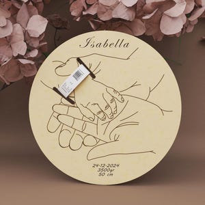 May include: A round wooden plaque with a hand-drawn illustration of hands holding a baby's hand. The name "Isabella" is at the top. The date 24-12-2024, weight 3500gr, and height 50 cm are at the bottom.