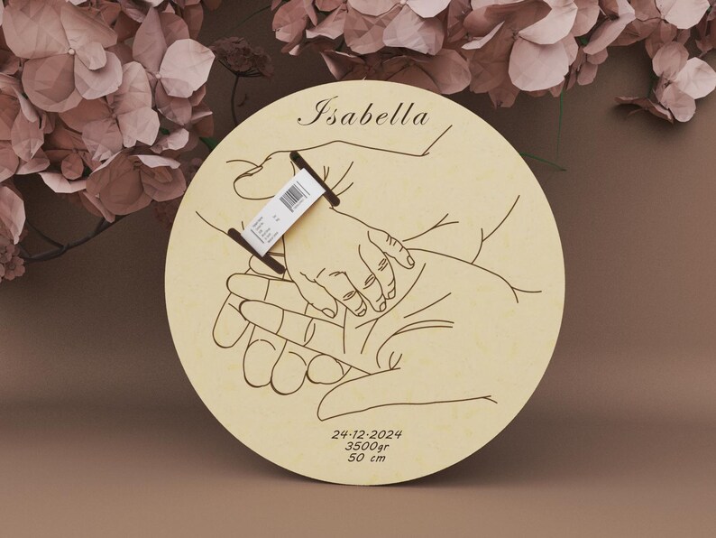 May include: A round wooden plaque with a hand-drawn illustration of hands holding a baby's hand. The name "Isabella" is at the top. The date 24-12-2024, weight 3500gr, and height 50 cm are at the bottom.