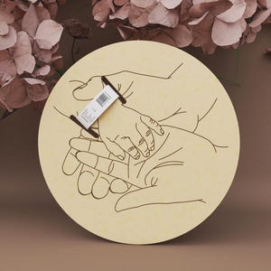 May include: A round wooden plaque with a line drawing of hands holding a baby's hand. The design is in black on a light beige background. A small tag is attached to the plaque. The background features dried flowers.