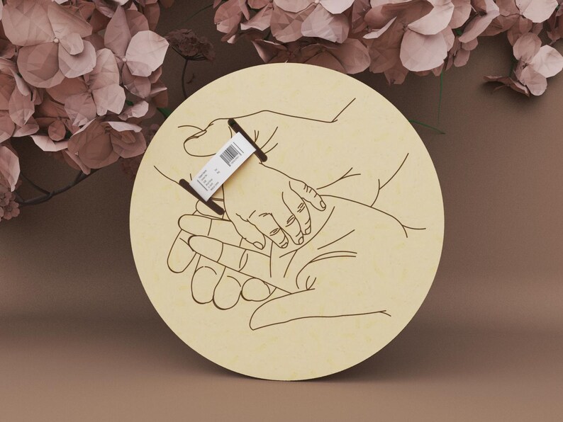 May include: A round wooden plaque with a line drawing of hands holding a baby's hand. The design is in black on a light beige background. A small tag is attached to the plaque. The background features dried flowers.