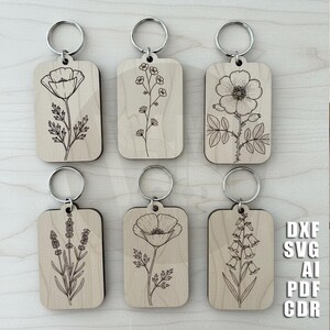 Puede incluir: Seis llaveros de madera con dise&ntilde;os florales grabados, incluyendo amapolas y lavanda. Cada llavero tiene un anillo plateado. Los llaveros son rectangulares con esquinas redondeadas. La imagen tambi&eacute;n incluye el texto "DXF SVG AI PDF CDR".
