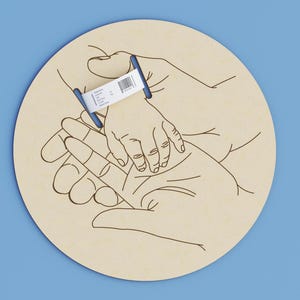 May include: A light beige wooden circle with a line drawing of hands holding a baby's hand. A blue and white hospital wristband is on the baby's wrist. The background is a solid light blue.