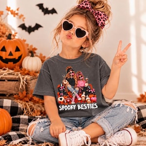 Puede incluir: Una camiseta gris carbón con un gráfico de dos figuras con rasgos esqueléticos, vistiendo ropa casual y accesorios, rodeadas de calabazas y hojas de otoño, con el texto Spooky Besties impreso en blanco en la parte inferior.