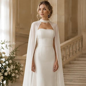 May include: A floor-length white gown featuring a strapless bodice and a high-neck sheer cape overlay. The dress has a fitted waistline and a long, flowing skirt that pools slightly at the floor, creating a clean and structured silhouette.