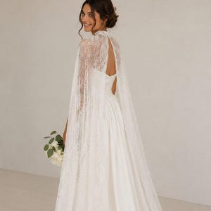 May include: A floor-length white wedding dress featuring a high-neck lace cape overlay. The cape is crafted from intricate floral lace, draping over the shoulders and back, while the dress underneath has a smooth, solid white fabric skirt.