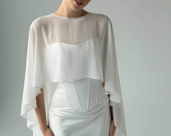May include: A white floor-length gown featuring a structured corset-style bodice with vertical seams and a sheer, lightweight cape overlay that drapes over the shoulders and arms, creating a layered silhouette with a clean, solid finish.
