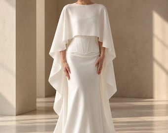 May include: A floor-length white gown featuring a fitted bodice with corset-style boning details and a wide, flowing cape overlay that drapes over the shoulders. The dress has a high neckline and a long, sweeping skirt that pools on the floor.