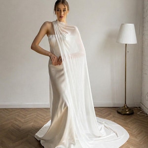 May include: A white bridal gown with a flowing cape. The dress has a simple, elegant design with a fitted bodice and a long train. The cape is sheer and drapes over the shoulders. A floor lamp with a white shade is in the background. The dress is made of a soft, silky fabric.