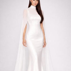 May include: A floor-length white satin gown featuring a fitted silhouette, a square neckline, and a sheer high-neck overlay with long, flowing cape-style sleeves that extend to the floor, creating a dramatic and elegant appearance.