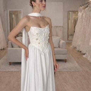 May include: A white wedding dress with a strapless bodice and a flowing skirt. The bodice is embellished with floral embroidery. The dress has a matching scarf. The dress is in a bridal shop.