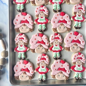 Könnte beinhalten: Ein Blech mit dekorierten Zuckerkeksen in Form der Figur Erdbeere. Jeder Keks hat eine rosa Haube mit roten Erdbeerakzenten, ein rotes Kleid und grün-weiß gestreifte Socken. Die Kekse sind auf einem Metallbackblech angeordnet.