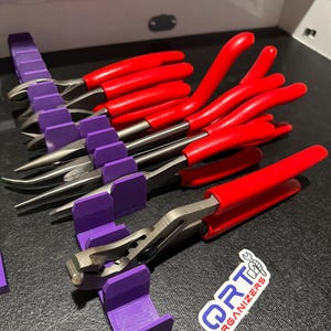 May include: A set of metal pliers with bright red handles arranged in a purple plastic organiser rack. The pliers are positioned in individual slots, and a white sticker with the blue and red text ORT ORGANIZERS is visible on the surface.