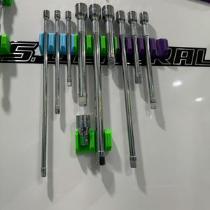 Puede incluir: Un conjunto de barras de extensión de metal plateado están organizadas en una pared magnética blanca con soportes de plástico verde, azul y morado. La pared tiene el texto "QRT Organizers" y "S. General Rail" impreso en ella.