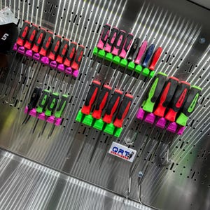 May include: A metal pegboard with a variety of screwdrivers and tools hanging on it. The tools are organised by colour and type. The pegboard is labelled "QRT ORGANISERS".