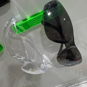 May include: Two pairs of safety glasses. One pair has clear lenses and a clear frame. The other pair has dark tinted lenses and a black frame. A green piece with the word "ORT" is visible.