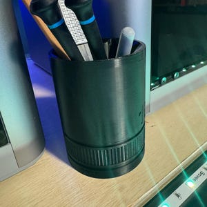 May include: A dark green cylindrical desk organizer, resembling a camera lens, holds tools. Inside are two black screwdrivers with blue accents, a pencil, and a white chalk stick. A metal ruler with measurements is also visible.