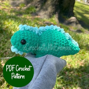 May include: A crochet pattern for a teal and light blue dinosaur. The pattern is available as a PDF download. The dinosaur is being held in a hand.