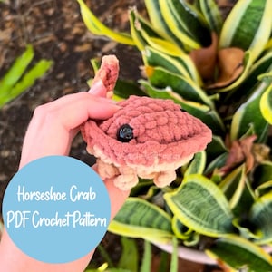 May include: A handmade, crocheted horseshoe crab in shades of dusty rose and cream. The crab has a textured body, small legs, and a black eye. The image includes a blue circle with the text "Horseshoe Crab PDF Crochet Pattern".