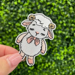 May include: Embroidered patch featuring a cartoon sheep with a pink bow, eyelashes, and small horns. The sheep is white with pink accents and a black outline. The patch is likely for clothing or accessories.