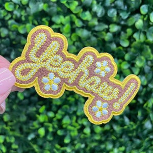 May include: Embroidered patch with the word "Hey" in yellow script, outlined in yellow and brown. Small white flower accents are scattered around the text. The patch is held against a green, leafy background.