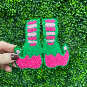 May include: A pair of festive elf shoes, perfect for holiday decorations. The shoes are green with pink and white striped socks and pink curled toes. The edges are outlined with glitter.