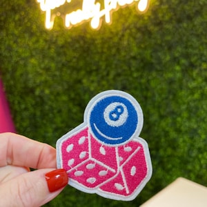 May include: Embroidered patch featuring a pink dice and a blue eight ball. The dice has white dots and the eight ball has a white number eight. The patch is white around the edges.