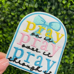 May include: Embroidered patch with the words "Pray on it, pray over it, pray through it" in yellow, pink, and blue. A light blue bow is at the top. The patch has a white background and a light blue border.