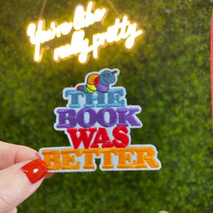 May include: Embroidered patch with the text "THE BOOK WAS BETTER" in purple, red, and orange. A colorful caterpillar sits above the text. The background includes the neon sign "You're like really pretty."