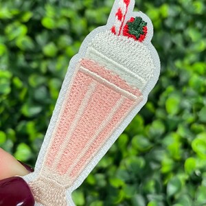 May include: Embroidered patch featuring a pink milkshake in a tall glass. The milkshake has white whipped cream, a red and white striped straw, and a green strawberry on top. The patch is white with a pink and beige color scheme.