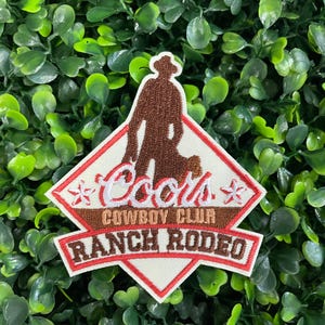 May include: Embroidered patch featuring a cowboy silhouette and the words "Coors Cowboy Club Ranch Rodeo." The patch has a diamond shape with a red border, white background, and brown and red text. Two white and red star-shaped designs are on either side of the word "Coors."
