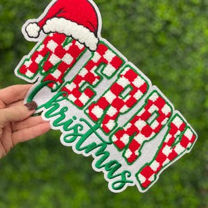 May include: A Christmas decoration with the words "Merry Christmas" in a playful, embroidered design. The letters are red and white checkered, with a green outline and a Santa hat on top. Ideal for festive holiday celebrations.