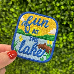 May include: Embroidered patch with a blue border and the words "fun at the lake" in blue script. The patch features a yellow sky with a sunset, green grass, and a brown bear.