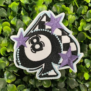 May include: Embroidered patch featuring a black and white eight ball design, with a checkered flag pattern and three purple stars. The patch has a white background and a light blue border.