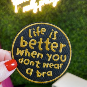 May include: A black circular patch with gold embroidered text that reads "life is better when you don't wear a bra". The patch has a gold border and is held by a hand with red nail polish.