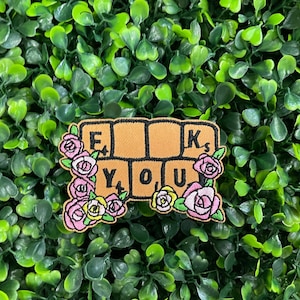 May include: A tan patch with the words "F**K YOU" spelled out in Scrabble-style tiles, surrounded by pink and yellow roses. The patch is set against a background of green foliage.