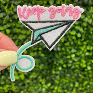 May include: Embroidered patch featuring a white paper airplane design with black trim and teal accents. The patch includes the pink text "Keep going" in a cursive font. The patch is held against a green background.