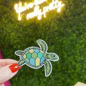 May include: Embroidered patch in the shape of a sea turtle, featuring a green, blue, and yellow geometric design. The patch is held in front of a green background with the neon text "You're really pretty".