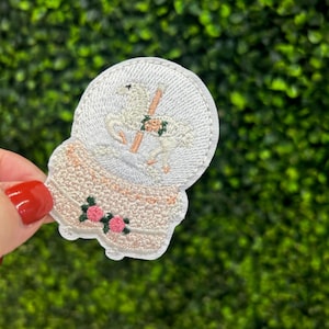 May include: Embroidered patch featuring a snow globe design. The patch depicts a carousel horse inside the globe, with a pink and green floral design at the base. The patch is white, with tan, pink, and green accents.