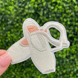 May include: Embroidered patch of two white ballet slippers with pink accents. The patch features detailed stitching and a delicate design, perfect for adding to clothing or accessories. The background is a blurred green.