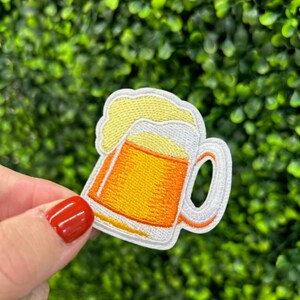 May include: Embroidered patch of a beer mug with yellow foam and orange beer. The mug has a white handle and outline. The patch is held in front of a green leafy background.