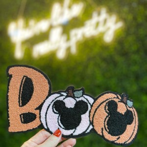 May include: A Halloween-themed patch with the word "Boo" in orange, followed by two pumpkin-shaped designs. The pumpkins feature Mickey Mouse silhouettes in black and are in shades of white and orange. The patch is held by a hand.