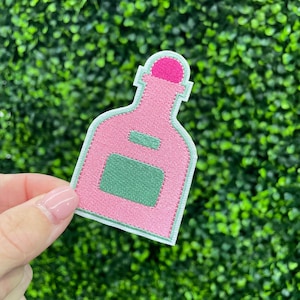 May include: Embroidered patch in the shape of a pink bottle with a green rectangular label and a pink cap. The patch has a light green border and is set against a green background.