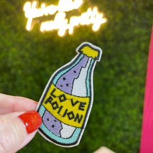 May include: Embroidered patch in the shape of a potion bottle. The bottle is light purple with a yellow label that reads "LOVE POTION" in black letters. The patch has a teal outline and a yellow cap.