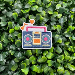 May include: Embroidered patch featuring a retro boombox design. The patch has a blue background with pink, orange, and white accents. A musical note and heart are above the boombox. The patch is set against a backdrop of green foliage.