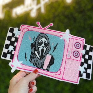 May include: Pink embroidered patch in the shape of a retro television. The screen displays a Scream mask character with lightning bolts. The patch has a black and white checkered pattern on the sides and white star accents.