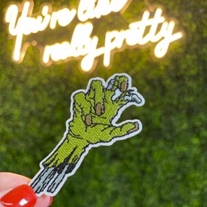 May include: A green zombie hand patch with a white border. The patch is held up in front of a green background with the words "You're like really pretty" in a cursive font.